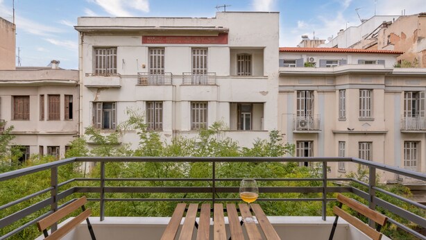 Un balcón con un banco de madera y una copa de vino, con vistas a un edificio con varias ventanas y balcones.