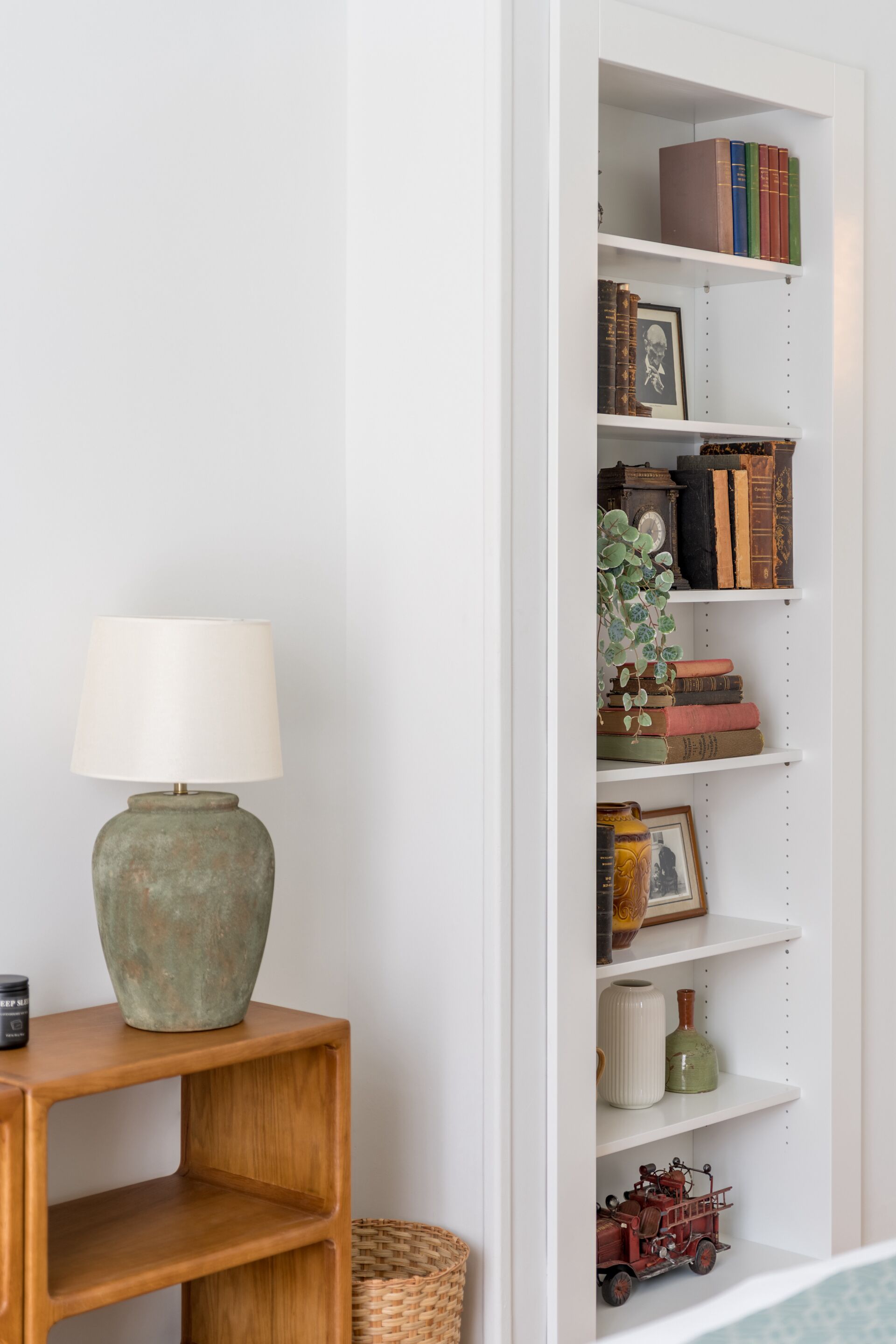 A bookshelf with books and decorative items, a lamp on a wooden side table, and a wicker basket.