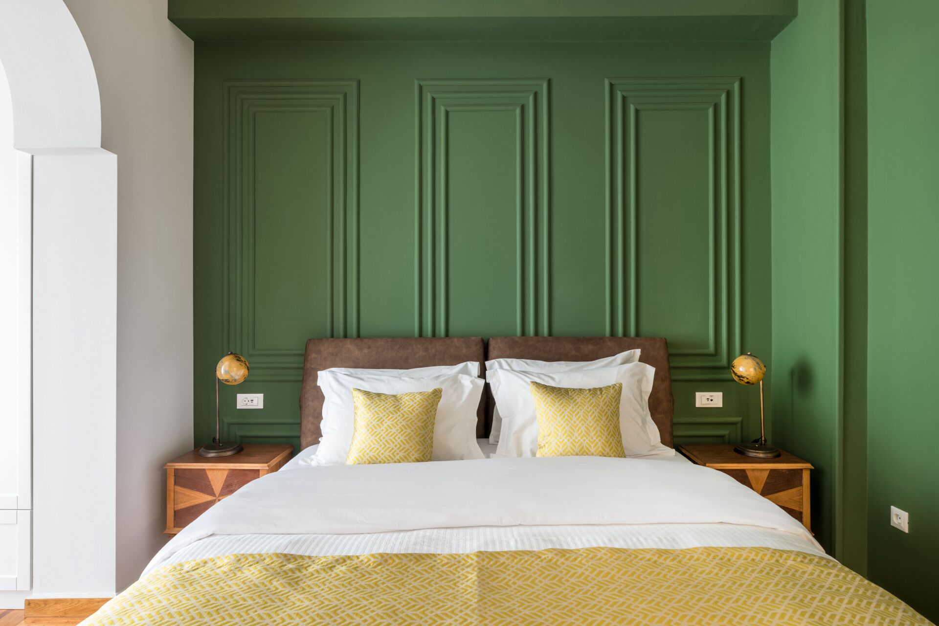Una cama bien tendida con ropa de cama blanca y amarilla estampada, dos lámparas de noche y una pared verde con paneles.