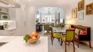 Una cocina moderna con armarios blancos, un comedor con mesa y sillas de madera, y una sala de estar con sofá y una obra de arte en la pared.