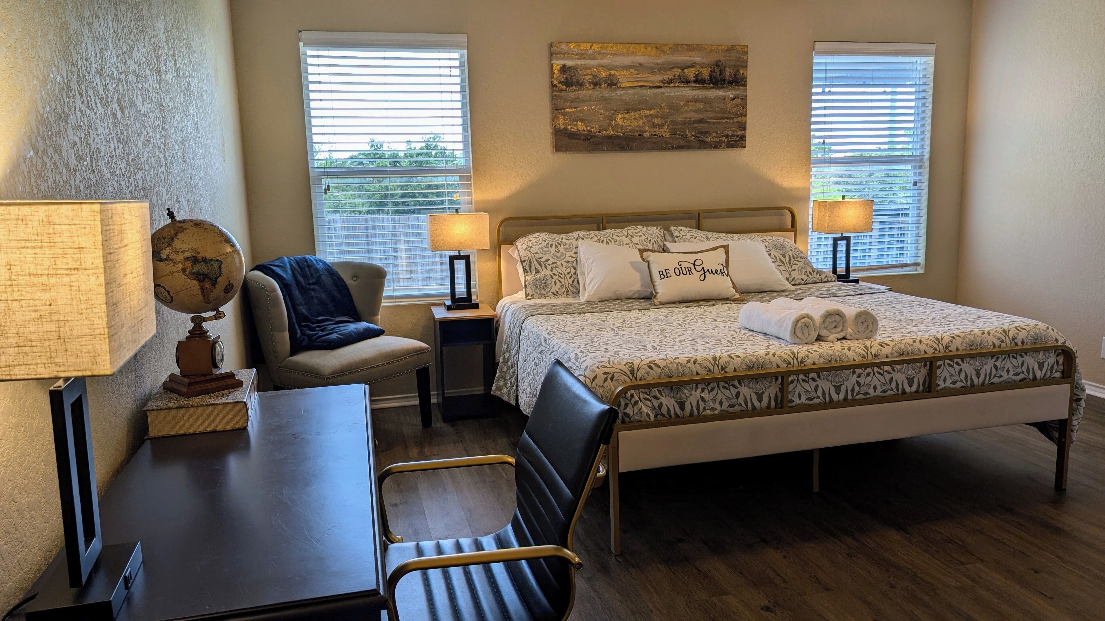A hotel room with a bed, two bedside lamps, a globe, a chair, a desk, and a window with blinds.
