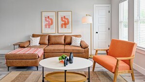 Een woonkamer met een bruine bank, een oranje fauteuil, een witte salontafel en twee abstracte kunstwerken in lijsten aan de muur.