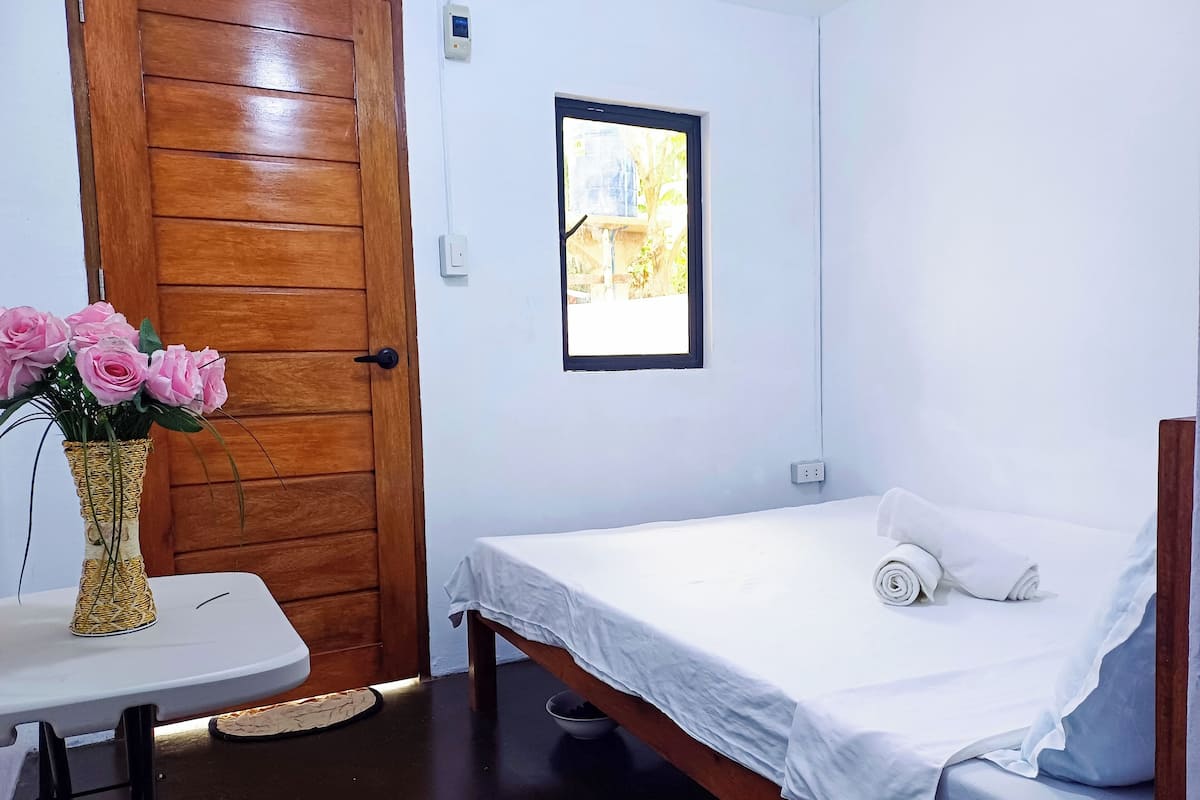 Una habitación pequeña y limpia, con puerta de madera, cama con ropa de cama blanca, una mesita con un florero de flores rosas y ventana con vista a follaje.