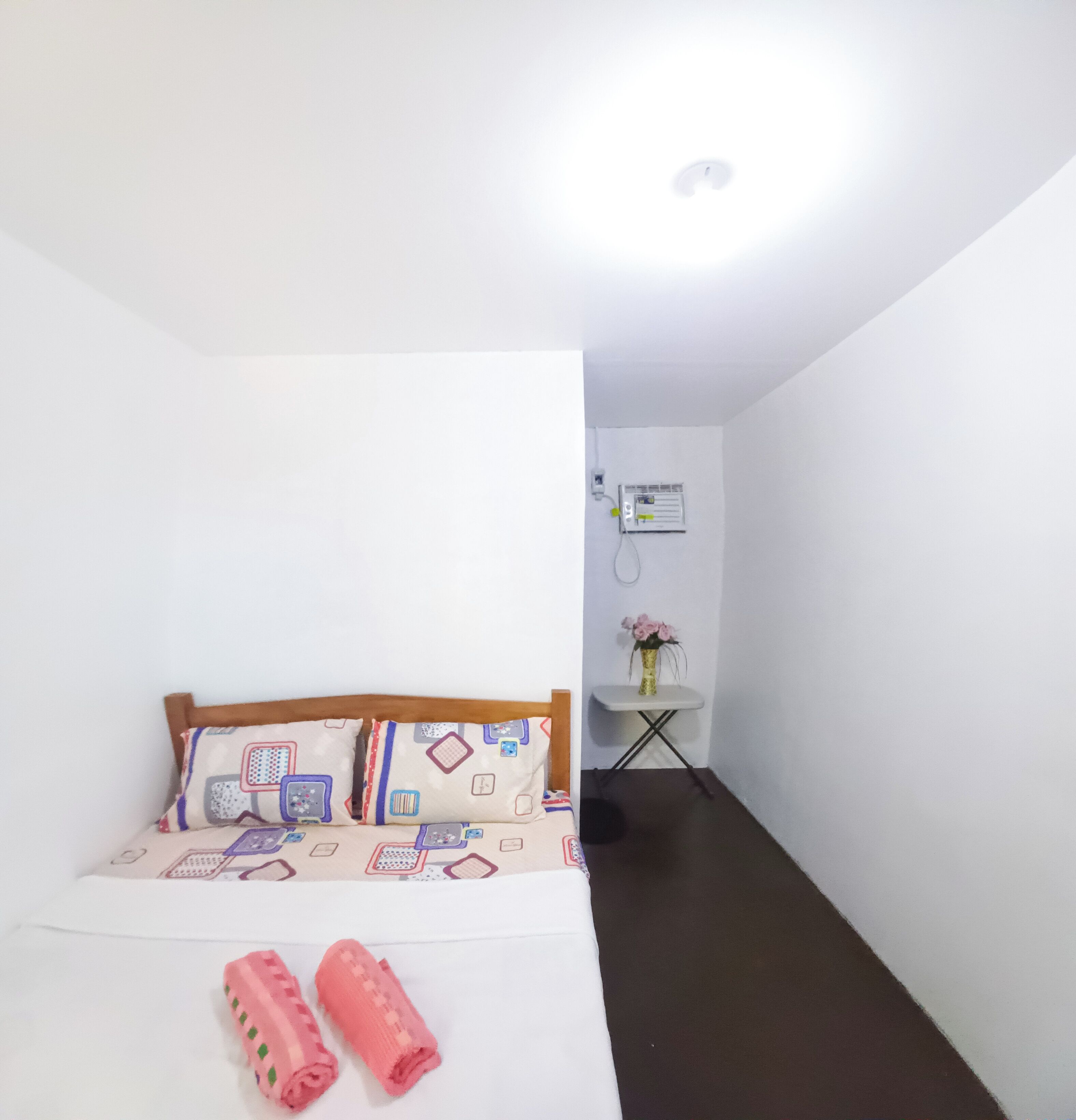 A small, clean room with a bed, pillows, a bedside table with a vase, and an air conditioning unit on the wall.