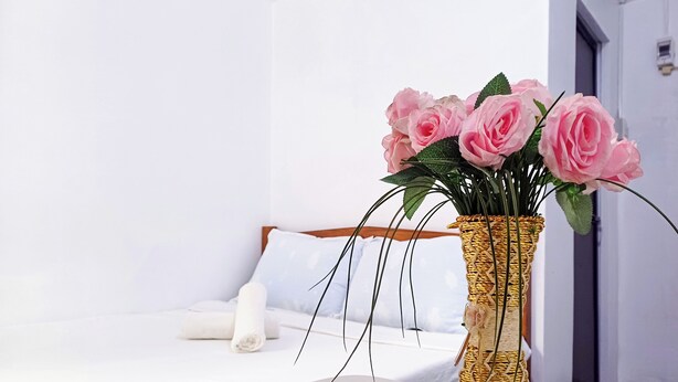 Un lit avec du linge de lit blanc, une serviette enroulée et un vase contenant des roses roses, posé sur une table ronde.