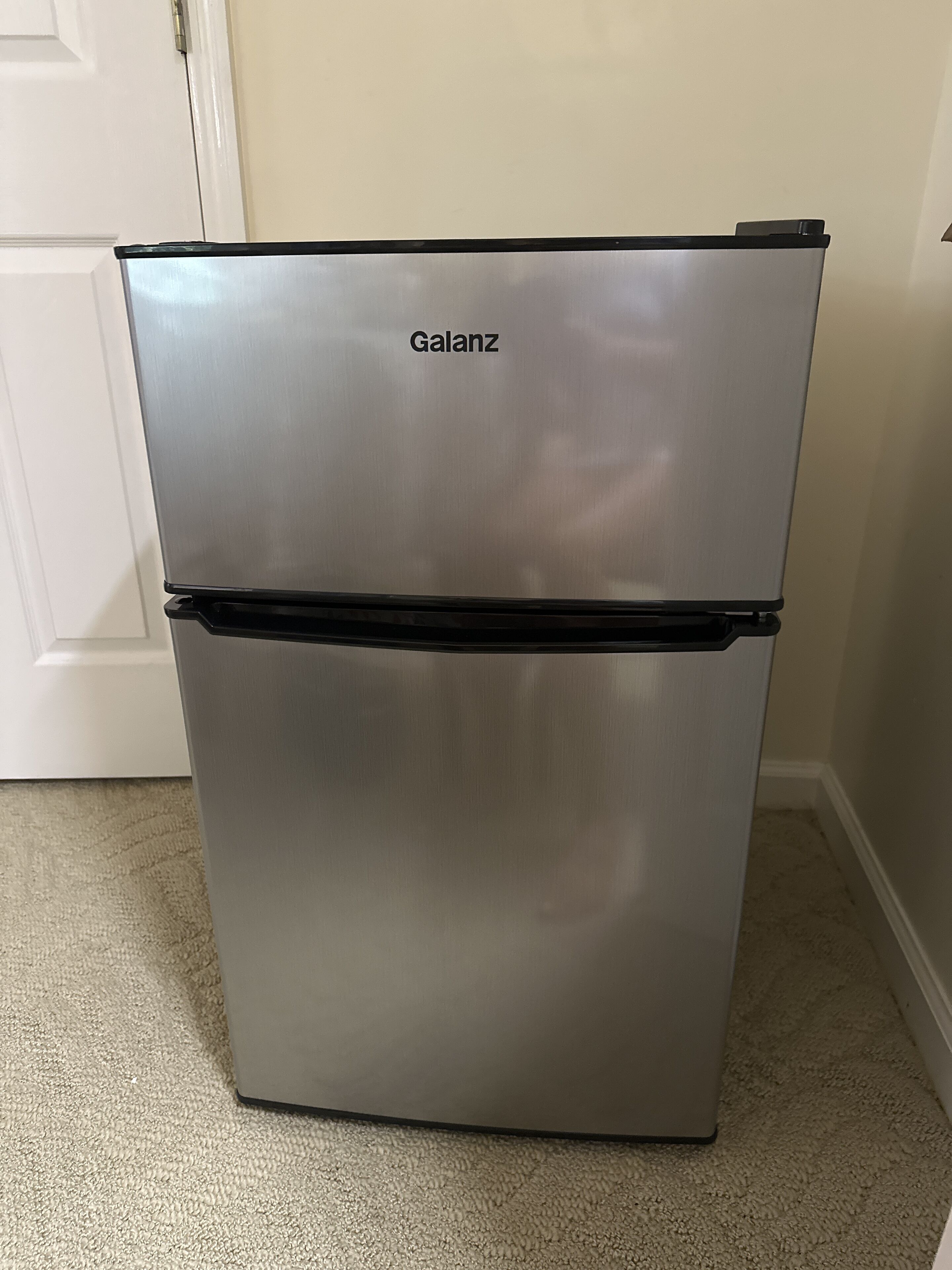 A stainless steel Galanz refrigerator with a black lower compartment, placed in a room with beige carpet and white walls.