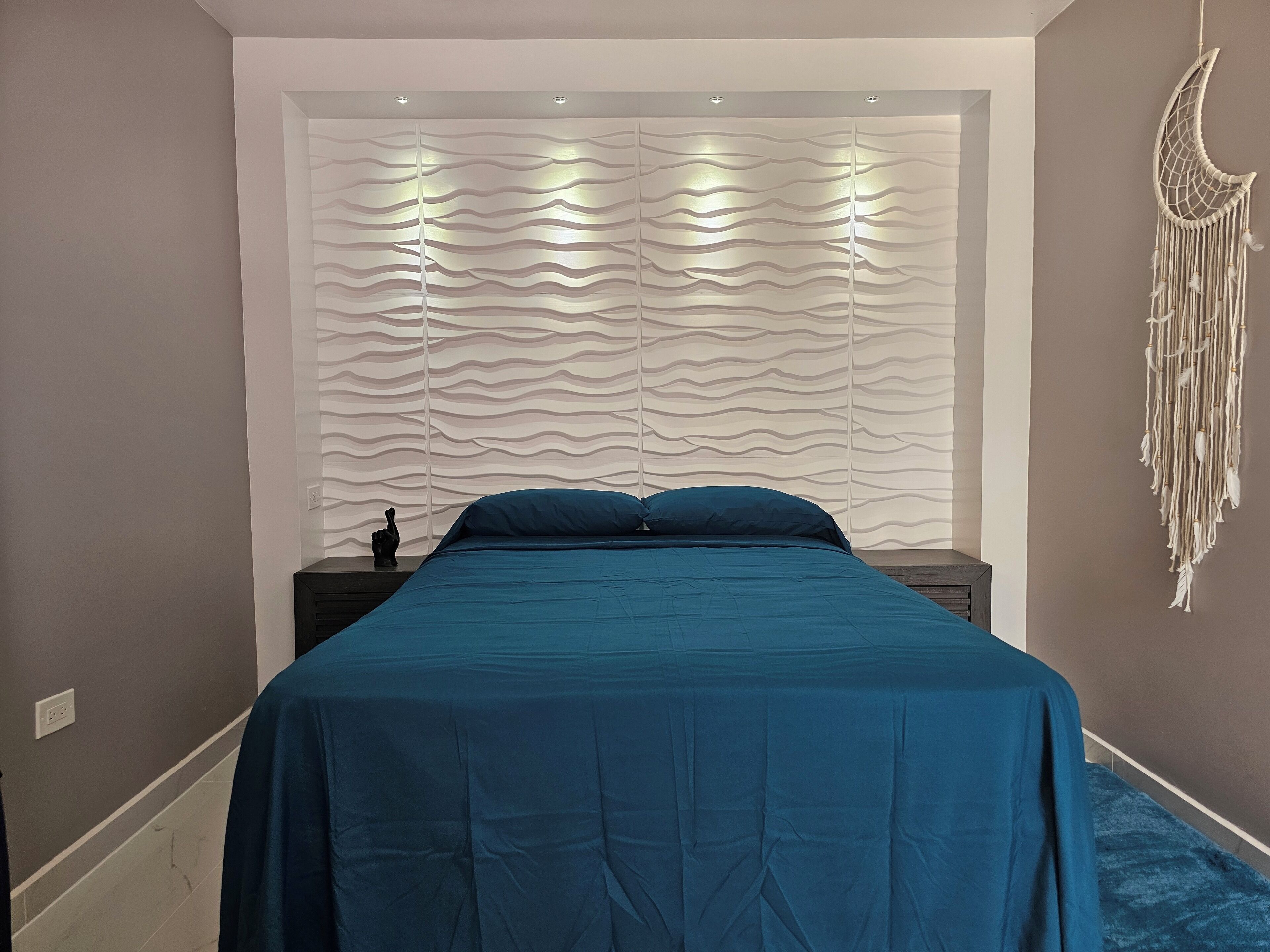 Een slaapkamer met een bed, een nachtkastje, een decoratief object aan de muur en een stopcontact.
