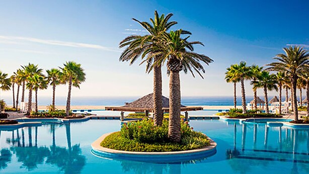 Zona de piscina en un resort con palmeras y vista al océano.