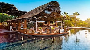 Un restaurante frente al mar con terraza cubierta, piscina y palmeras al fondo.