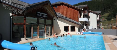 A swimming pool area with people swimming and lounging, a wooden building with a sign, and a mountain in the background.