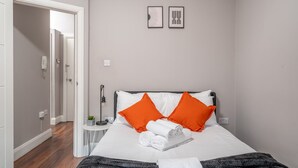 A neatly made bed with white and orange pillows, a small bedside table with a lamp, and a wall-mounted light switch.