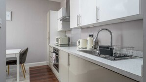 A compact kitchen with white cabinets, a stainless steel sink, a microwave, a toaster, and a kettle. There is a small dining table with a chair and a white rug on the floor.