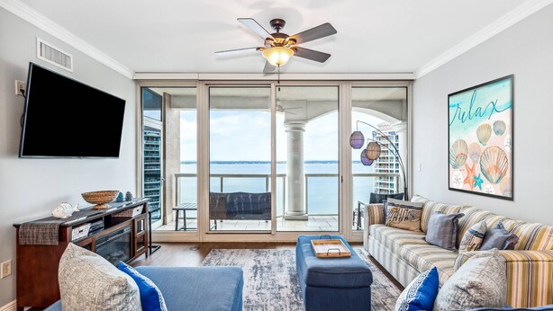 A spacious living room with a large flat-screen TV, a ceiling fan, and a view of the ocean through floor-to-ceiling glass doors.
