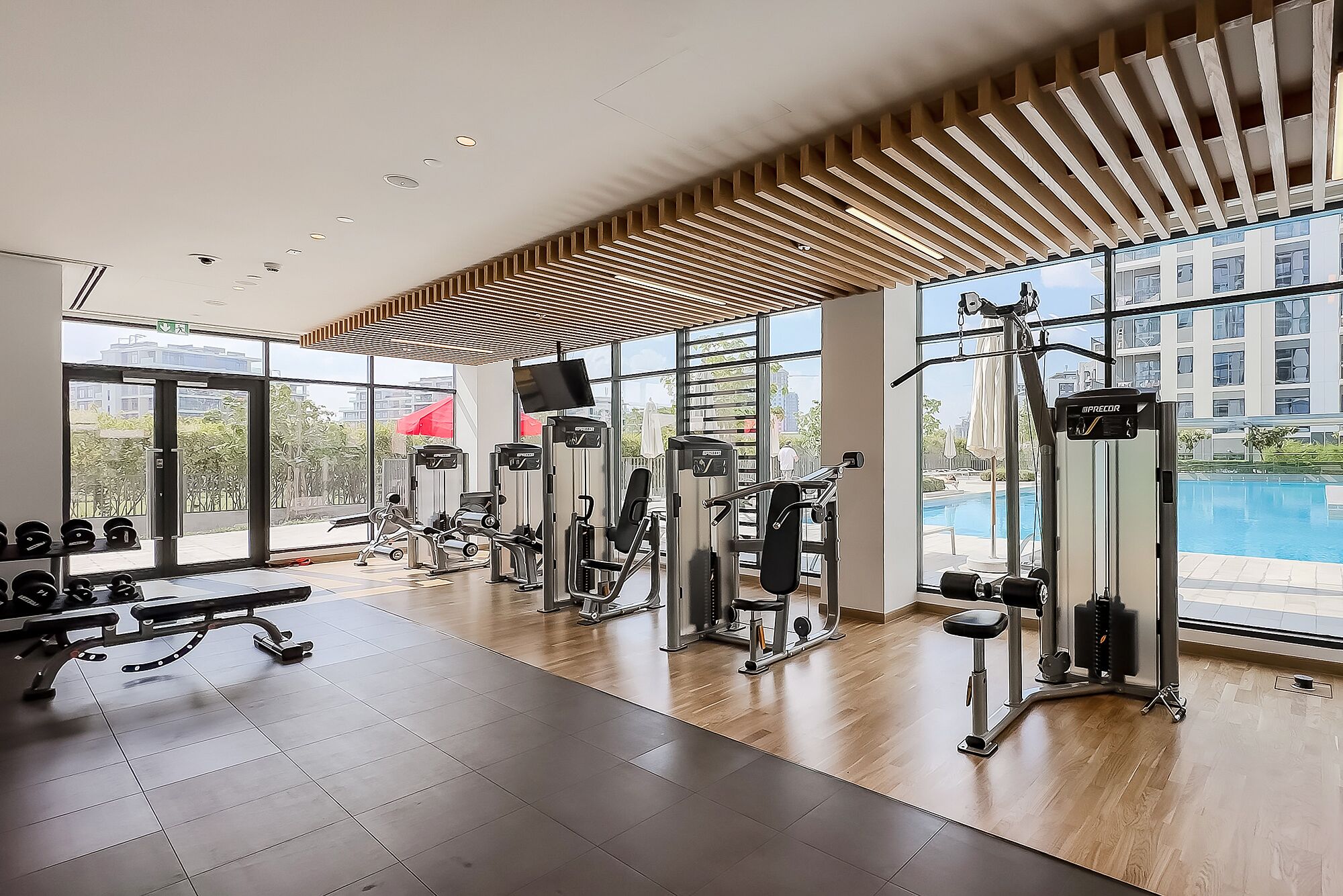 Et moderne fitnesscenter med diverse træningsmaskiner, store vinduer og udsigt til en pool.