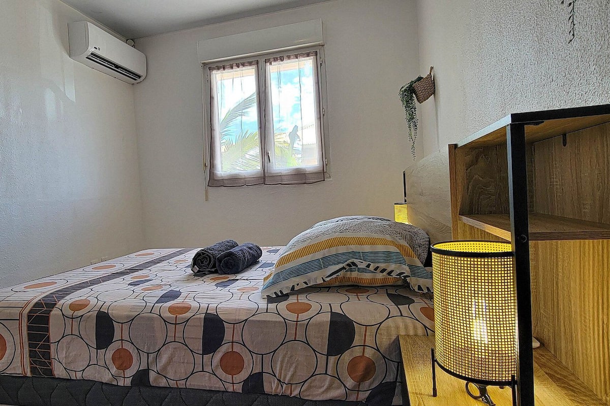 A bedroom with a bed, a bedside lamp, a window, and an air conditioning unit.