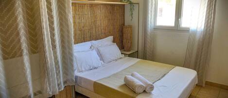 Una cama bien tendida con sábanas blancas y toallas enrolladas, un cabecero de bambú y una ventana con cortinas.