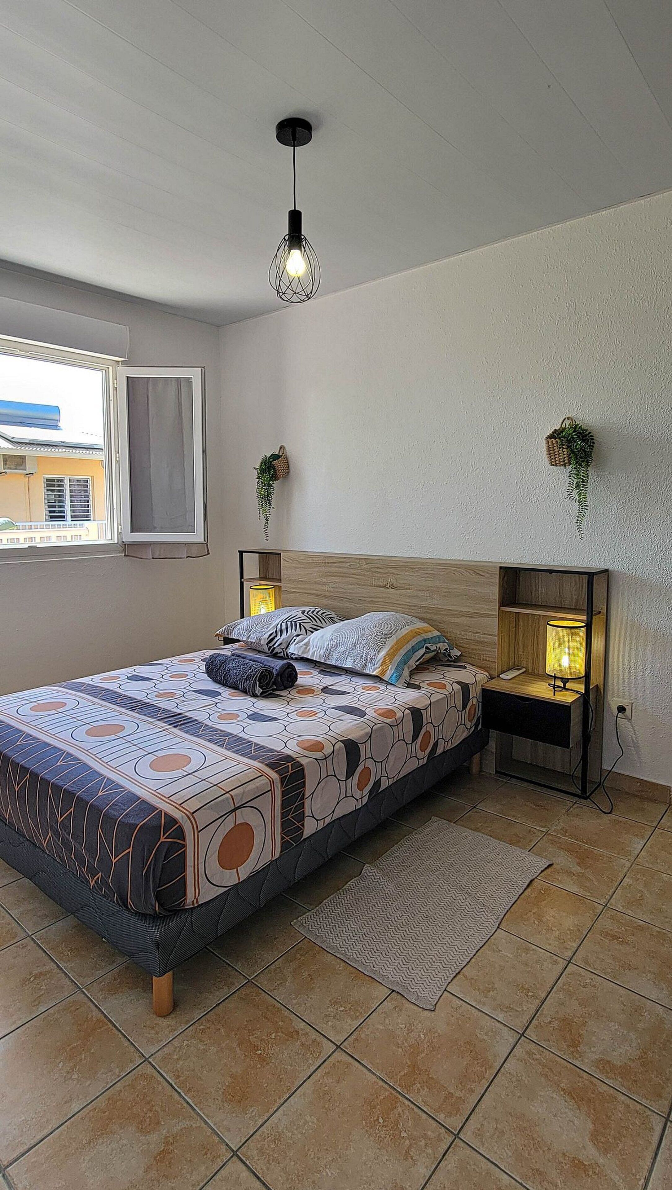 A bedroom with a bed, two bedside tables, a hanging light fixture, a window, and a view of the outside.