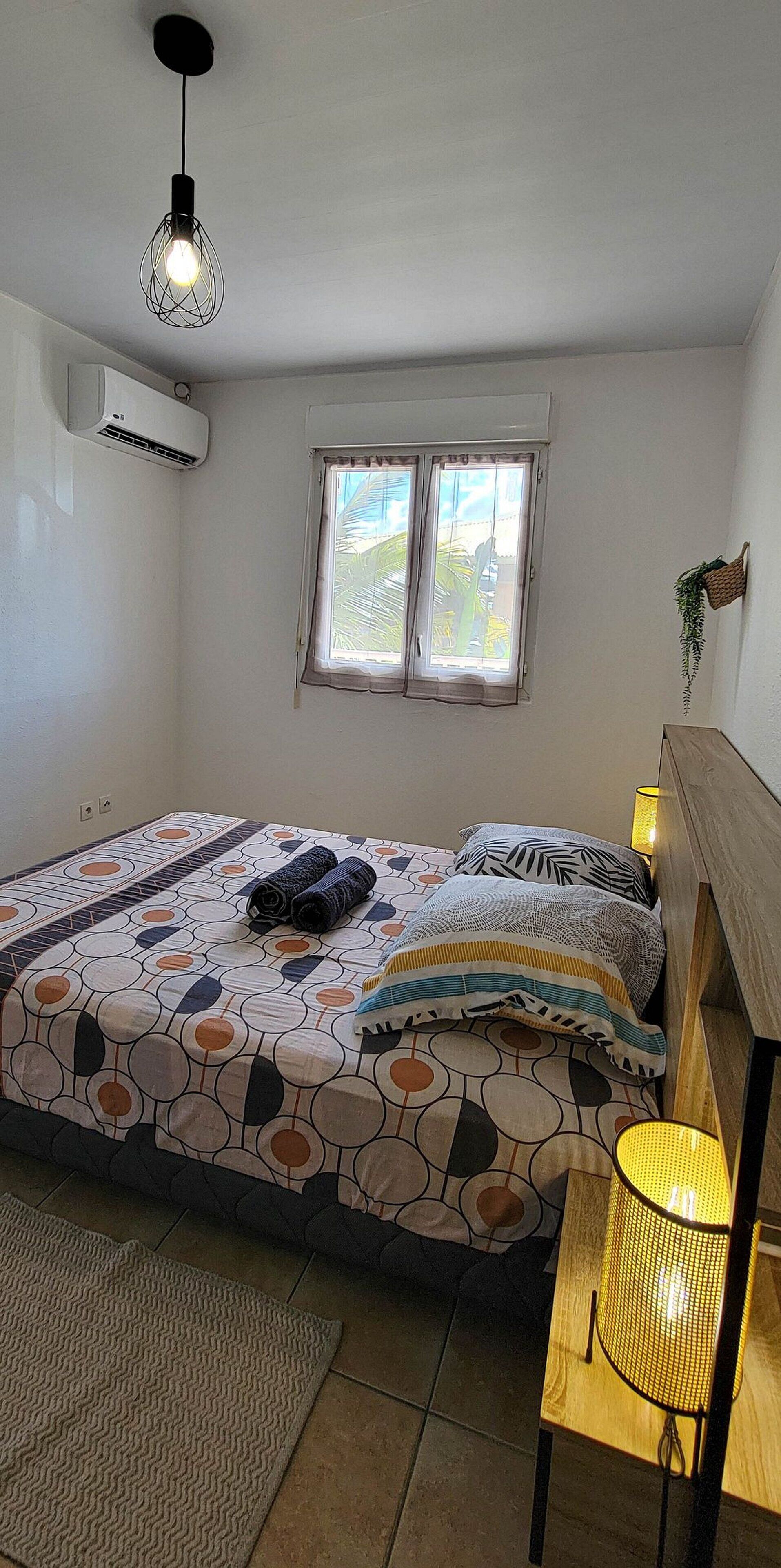 A bedroom with a bed, a window, an air conditioner, and a lamp.