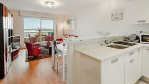 A modern kitchen with white cabinetry, a dining area with red chairs, and a view of the sea through large windows. - Terrasses d'Emeraude by Interhome (Dinard)