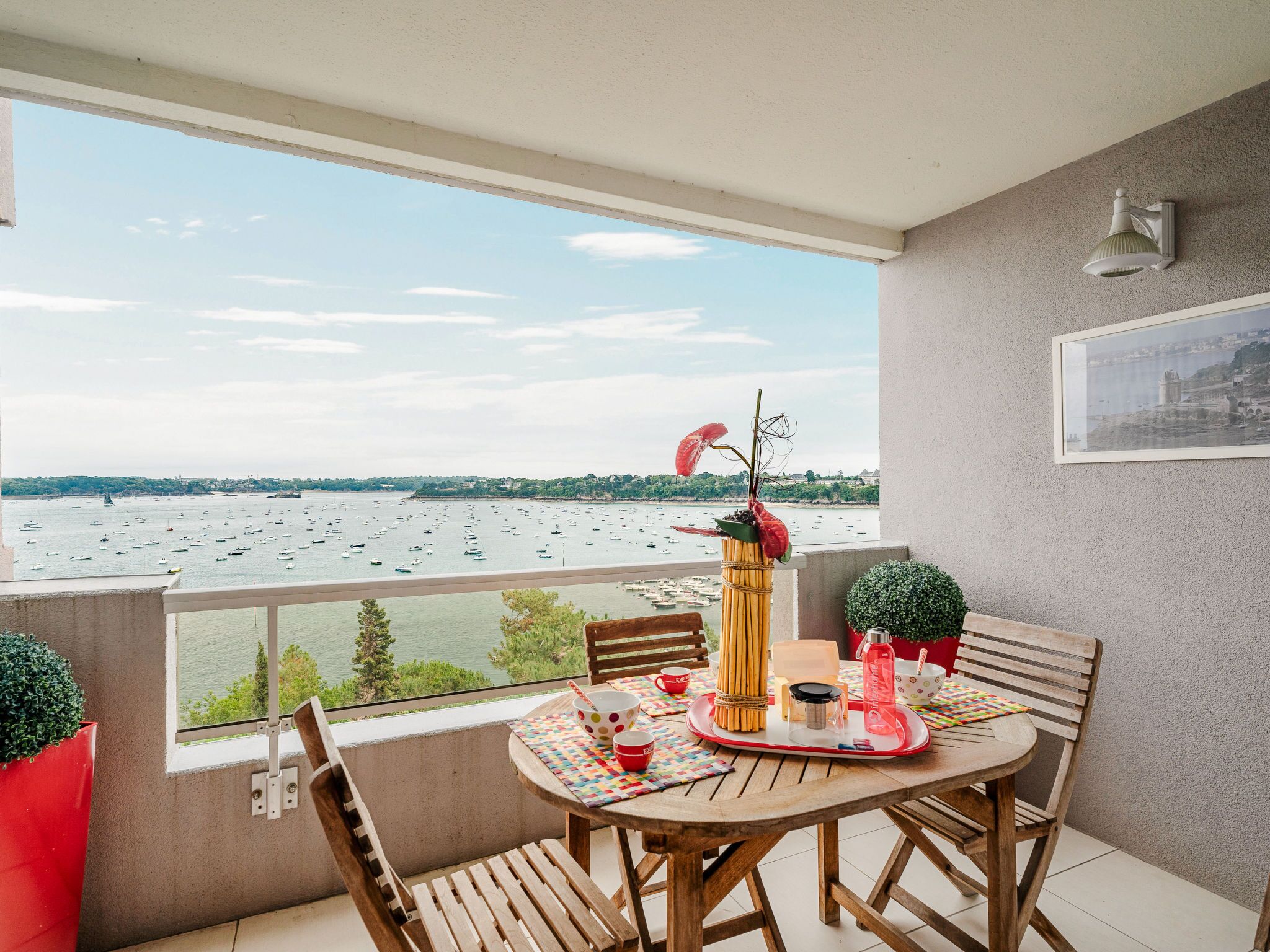 A balcony with a wooden table, chairs, a small plant, and a view of boats on the water.