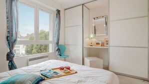 A bedroom with a bed, a chair, a bedside table with a lamp, a mirror, and a window with curtains. - Terrasses d'Emeraude by Interhome (Dinard)