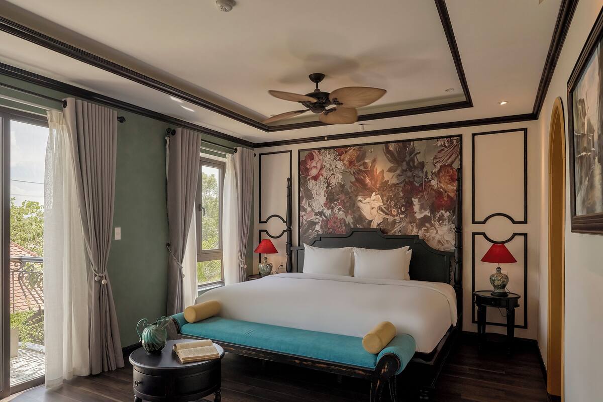 Una cama grande con cabecera, un ventilador de techo, una ventana con cortinas y una pared con un mural floral.