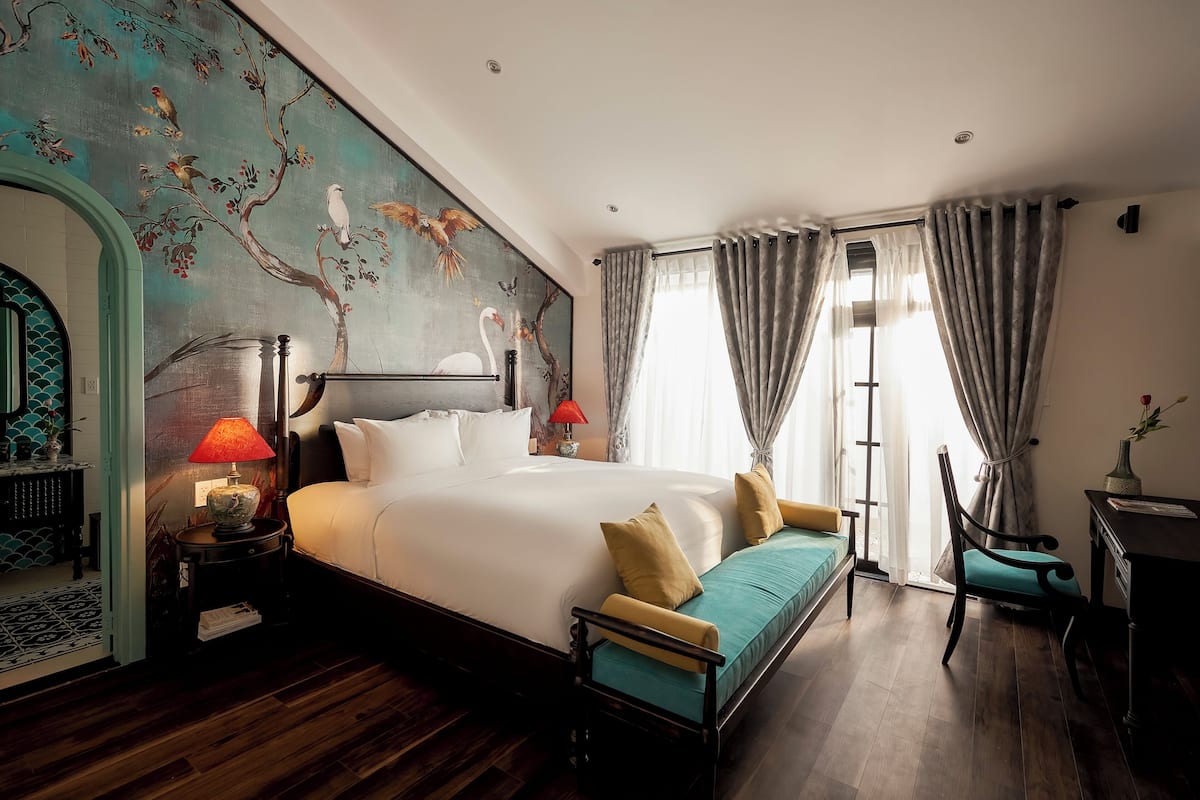 Habitación de hotel con una cama grande, un sofá, un escritorio y una silla. Hay una ventana con cortinas, una pared con un mural y una lámpara en una mesita de noche.