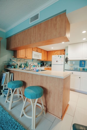 A kitchen with wooden cabinets, a central island with stools, a microwave, and a refrigerator. - Errol by the Sea unit 205- 1 BD - Tide & Joy (New Smyrna Beach)