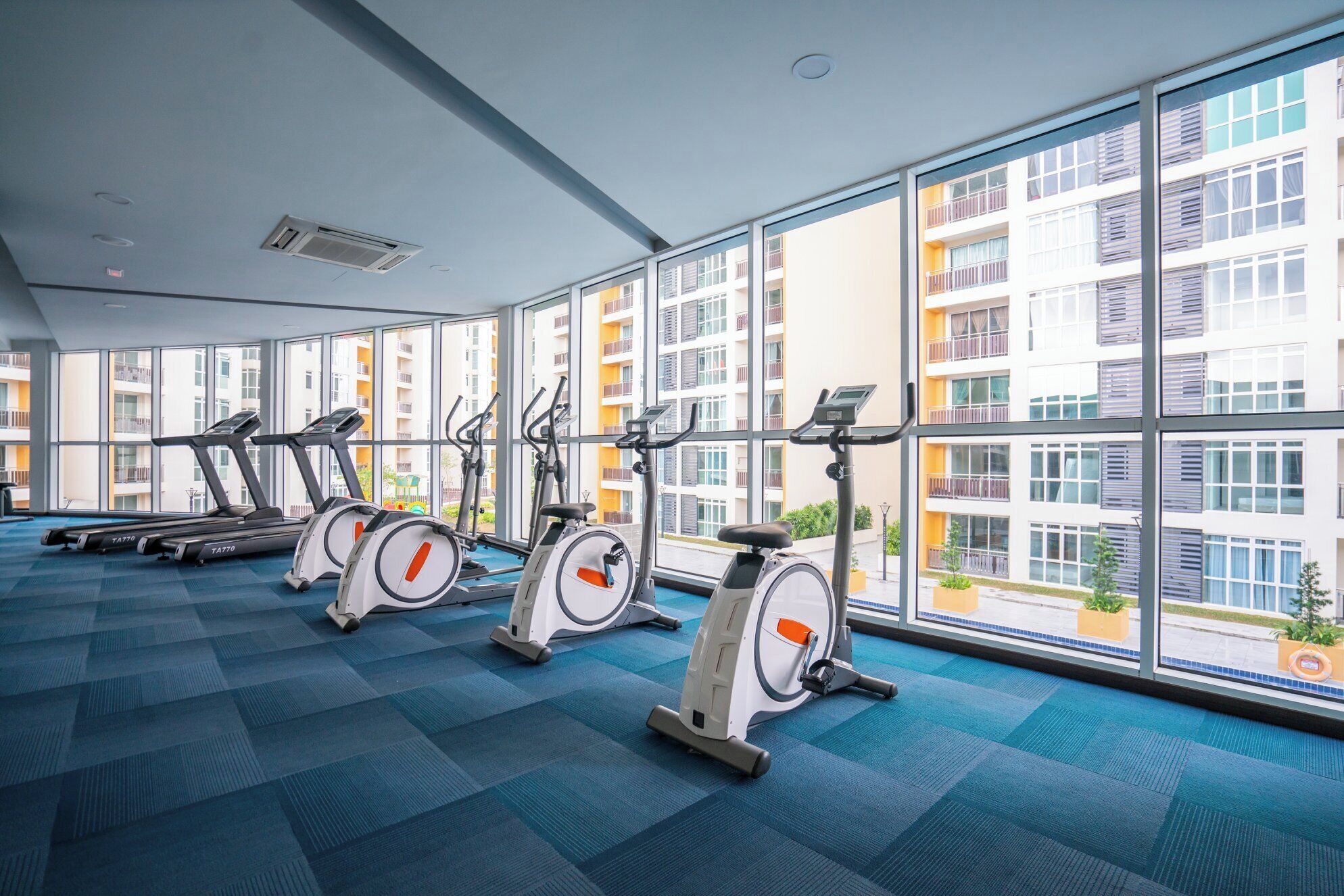 Une salle de sport équipée de quatre vélos d’appartement et d’un tapis de course, dotée de grandes fenêtres et d’un sol recouvert d’une moquette bleue.