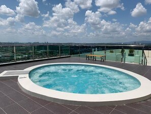 Una piscina en la azotea con un banco, rodeada por una baranda de cristal, con vistas a un paisaje urbano.