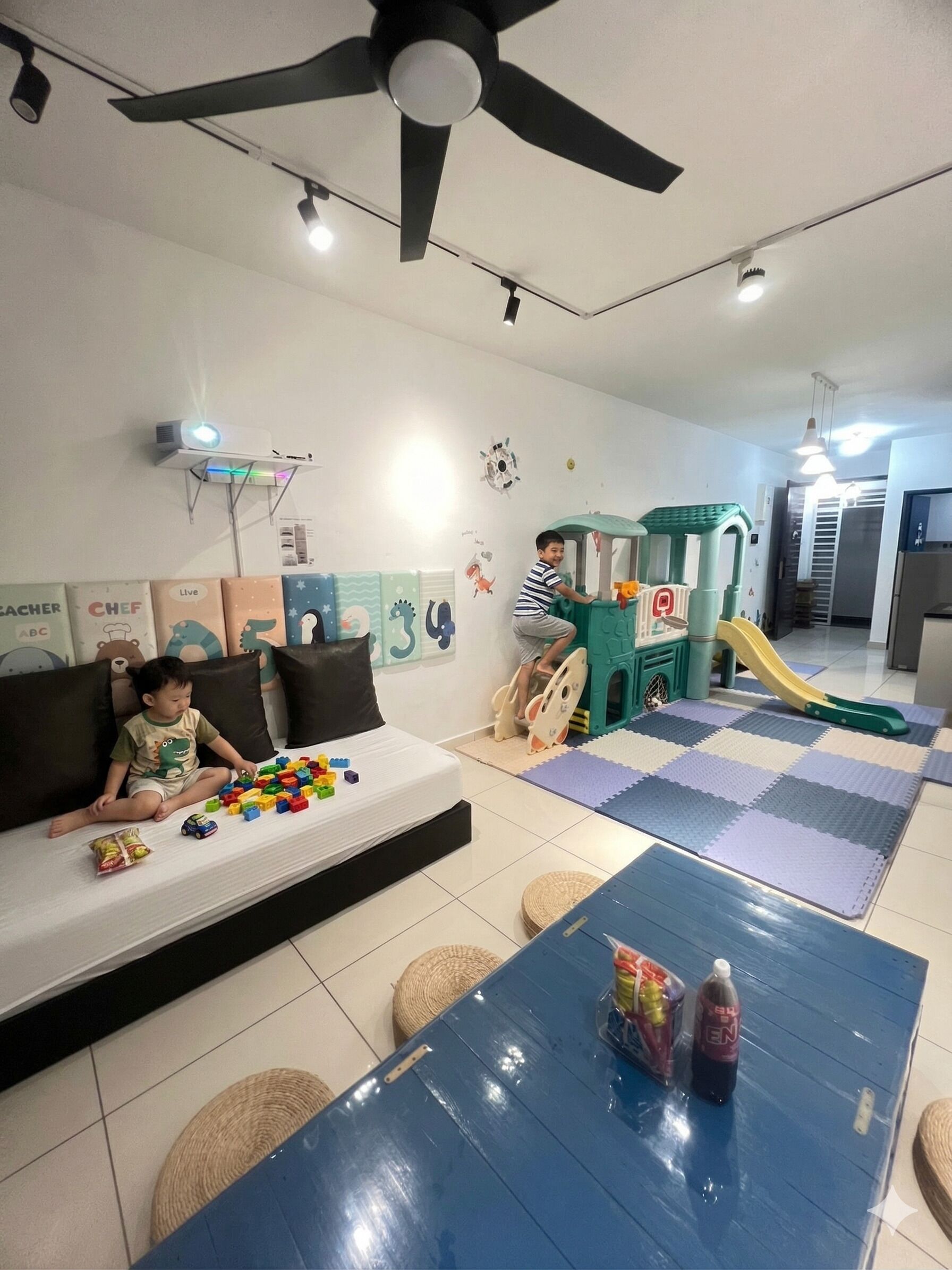 A living room with a sofa, play area, ceiling fan, projector, and play mats.
