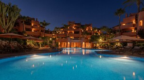 A swimming pool with lounge chairs and umbrellas, surrounded by multi-story buildings with balconies and palm trees. - The Signature Penthouse (Marbella)