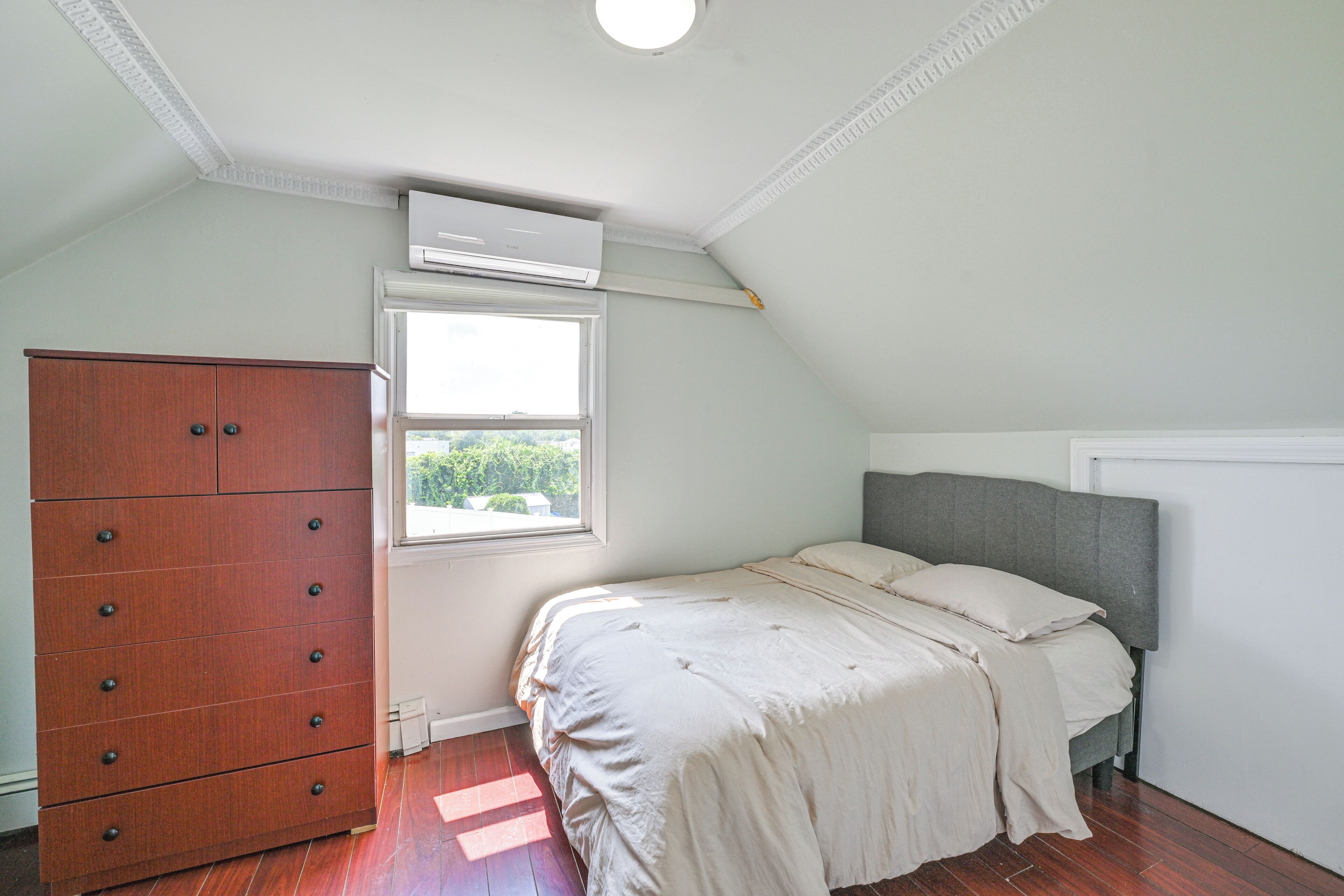 Une chambre avec une commode en bois, un lit doté d’une tête de lit grise et une fenêtre laissant entrer la lumière naturelle.