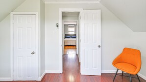 Una habitación con piso de madera, paredes blancas y una puerta blanca. Se observa una silla de color naranja brillante y una cama con almohadones azules al fondo.