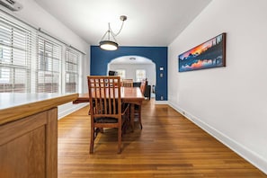 A dining area with a wooden table and chairs, a hanging light fixture, and a painting on the wall.