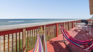 Eine Holzterrasse mit Blick auf Strand und Meer. Zwei Hängematten und ein Tisch mit Stühlen sind zu sehen.