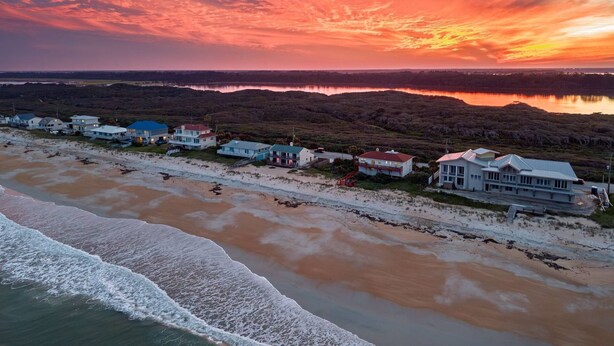 Eine Küstenlandschaft mit Häusern am Strand, freier Blick auf den Ozean und einem Sonnenuntergangshimmel.