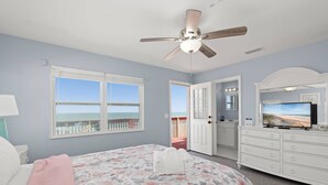 Ein Schlafzimmer mit Bett, Deckenventilator, Kommode, Fernseher und Fenster mit Blick auf den Strand.