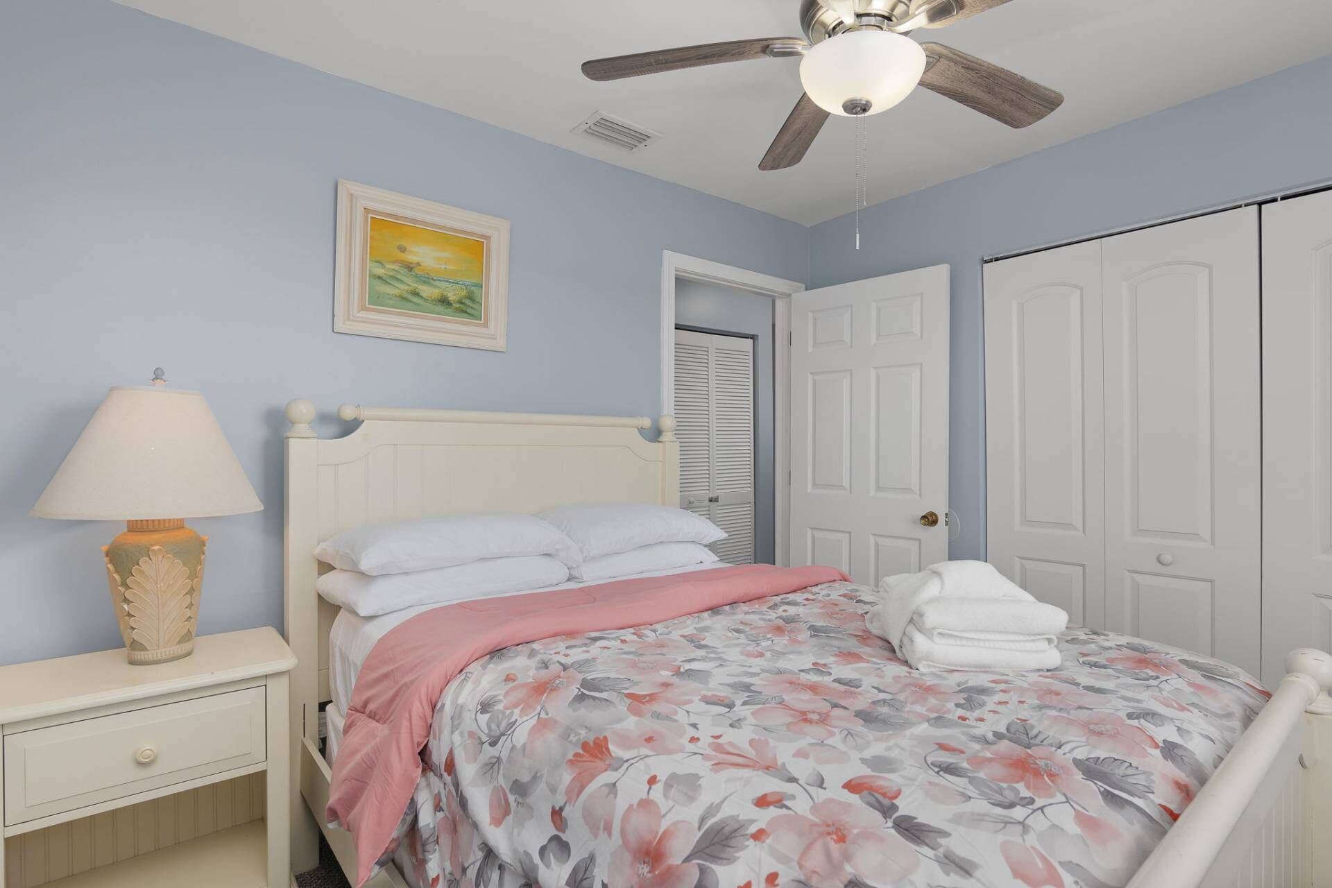 Quarto com cama, mesa de cabeceira, abajur, ventilador de teto e porta.
