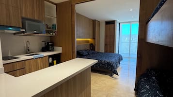 A modern kitchen with wooden cabinets, a microwave, and a coffee maker. There is a bed with a dark patterned bedspread in the background.