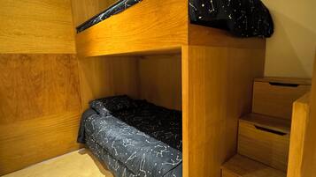 Una cama nido con estructura de madera y un colchón azul oscuro con diseños blancos. A la derecha hay un aparador de madera y una escalera que lleva a un nivel superior.