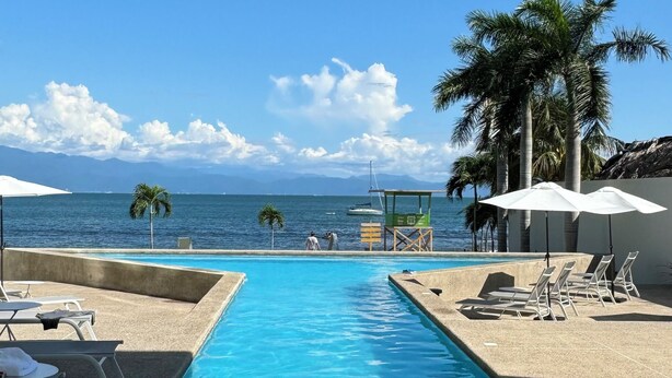 Una piscina con sillas de descanso y sombrillas, una torre de salvavidas y vistas al océano y a las montañas.