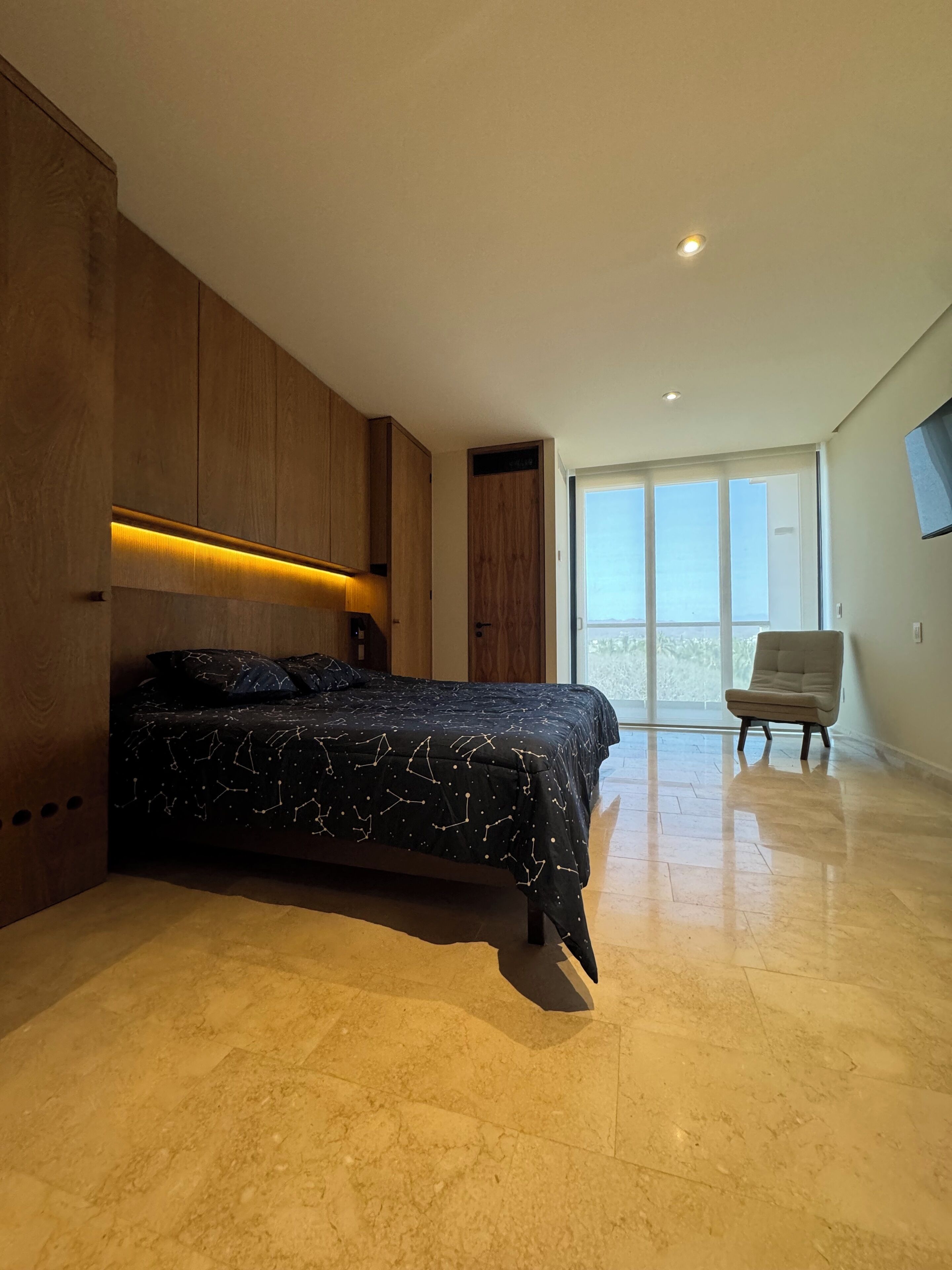 Un dormitorio moderno con una cama grande, muebles de madera, una silla y un amplio ventanal.