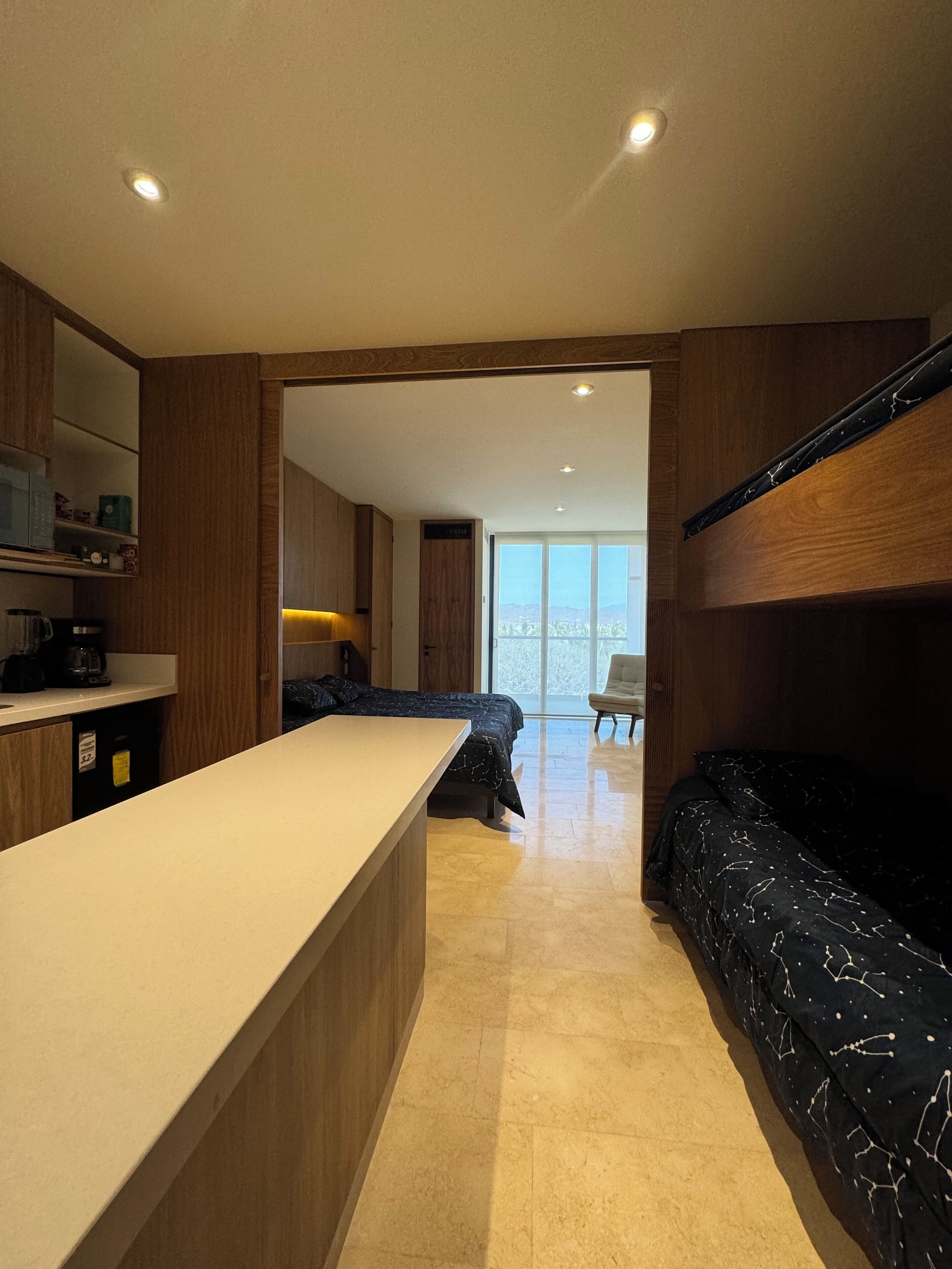 Una cocina moderna con una isla central, un área de comedor con un sofá negro estampado, y vistas a un balcón con el paisaje urbano.