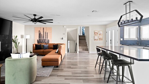 A modern living room with a sectional sofa, a green ottoman, and a ceiling fan. A kitchen area with bar stools and a backsplash is visible in the background. - Heated Pool, Hot Tub, Game Room & Sport Court (Glendale)