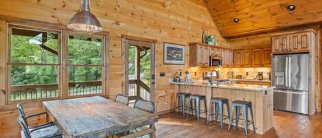 A wooden cabin interior with a dining area, kitchen, and large windows offering views of the outdoors.
