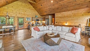 A spacious living room with a wooden ceiling, a large L-shaped sofa, a dining table, and a kitchen area visible through an open door. - Surreal Views~Outdoor Fireplace Pool Table&Hot Tub (Epworth)