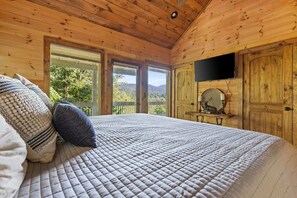 A wooden cabin bedroom with a large bed, a television, and a view of the forest.