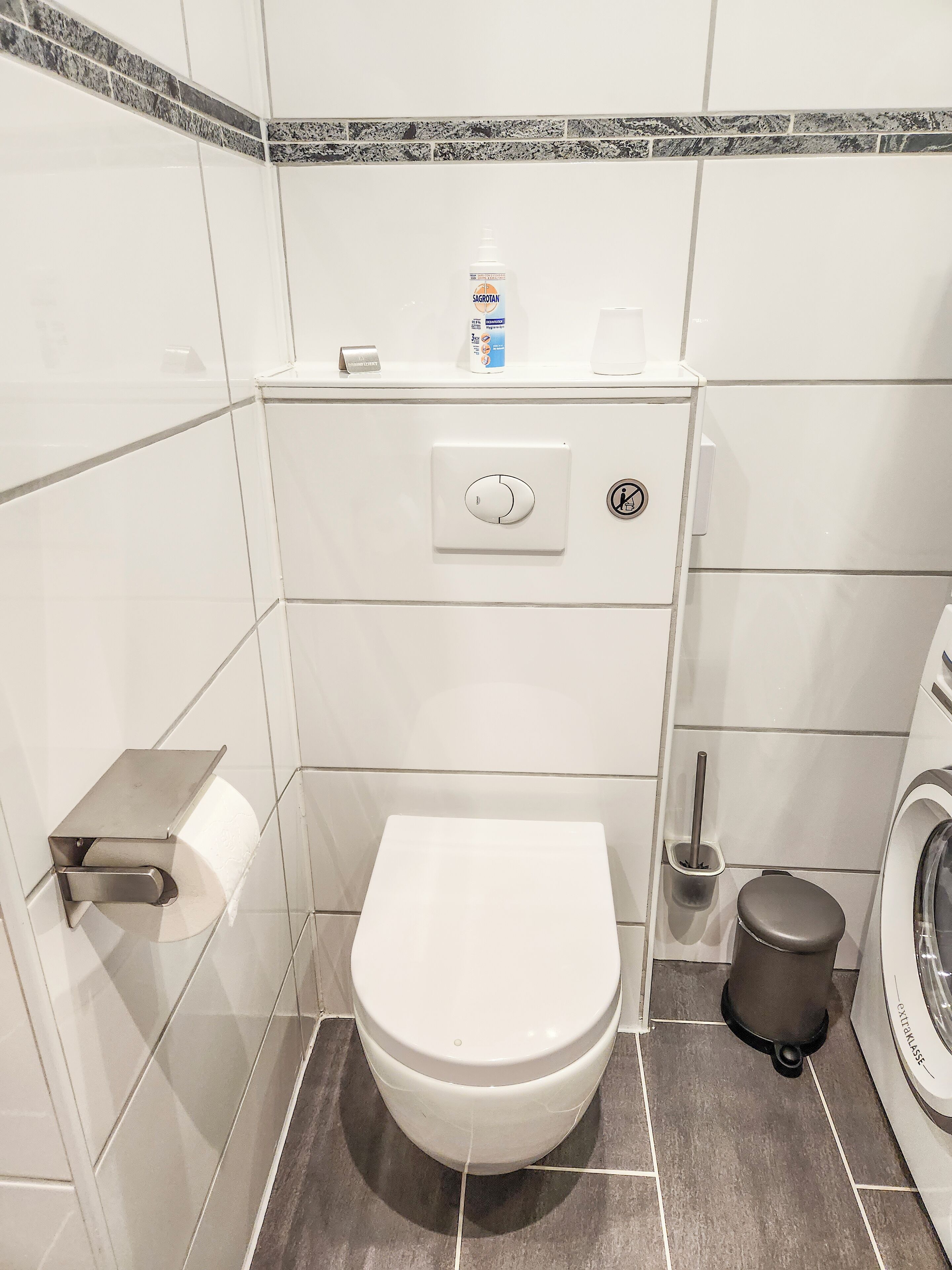 Een kleine badkamer met toilet, handdroger, papieren handdoekdispenser en wasmachine.