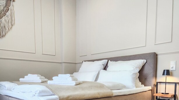 A large bed with multiple pillows and a blanket, a wooden floor, and a wall-mounted lamp. - Geli Homes L1 - 2x Kingsize Bett, 1 bis 4 Pers, Parkplatz, TV, Wifi, Kaffee, Tee (Lünen)